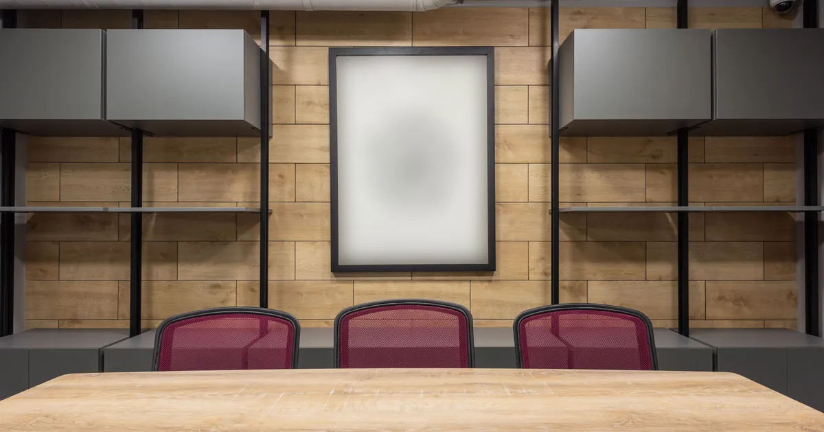 Sala de juntas vacía con mesa de madera clara y tres sillas color vino frente a un muro de madera con un cuadro en blanco
