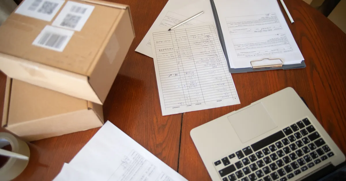 Cajas de envío etiquetadas junto a una hoja de registro de entregas, formatos en una tabla con clip y una laptop sobre una mesa