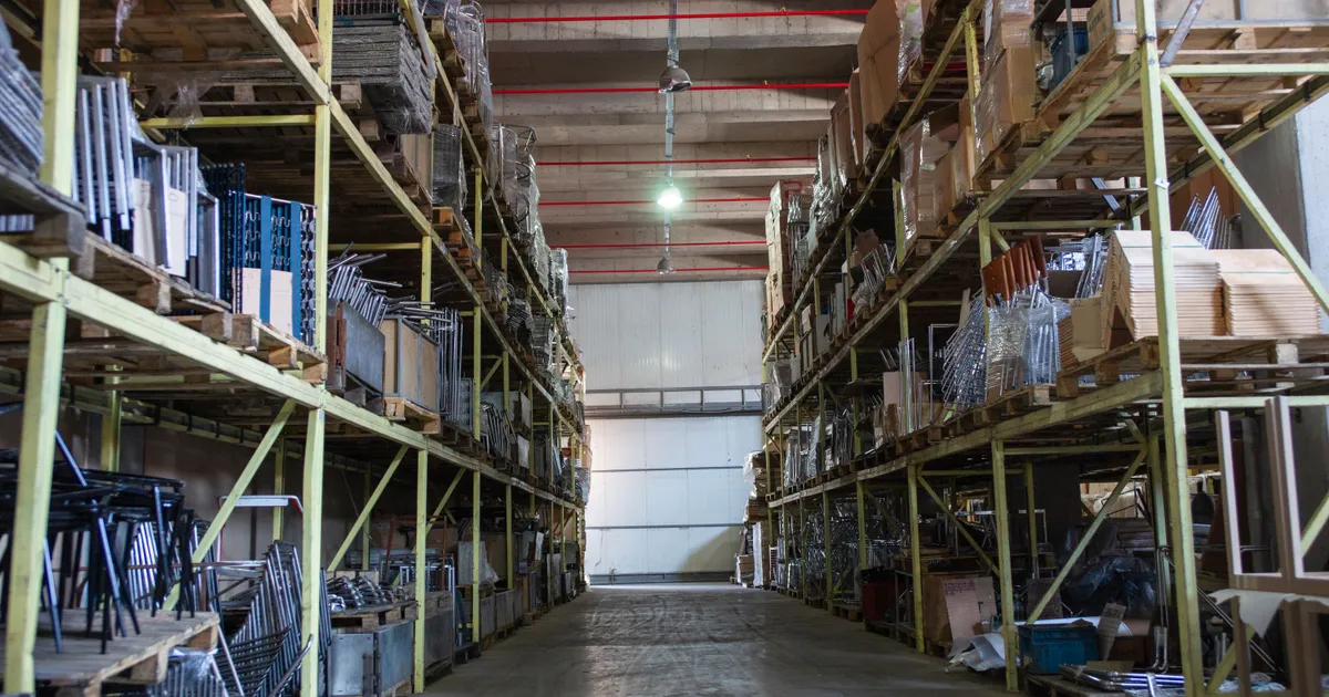 Pasillo de un almacén con estanterías metálicas cargadas de cajas, tarimas y estructuras de metal