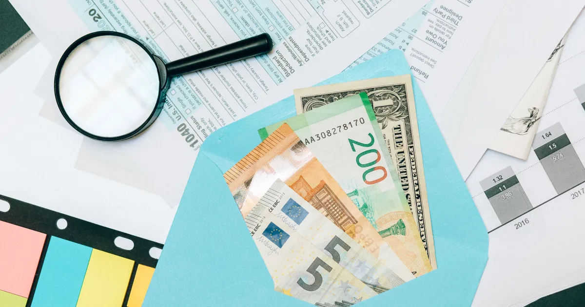 Sobre azul con billetes de euro y de dólar sobre formularios fiscales, junto a una lupa y notas adhesivas de colores